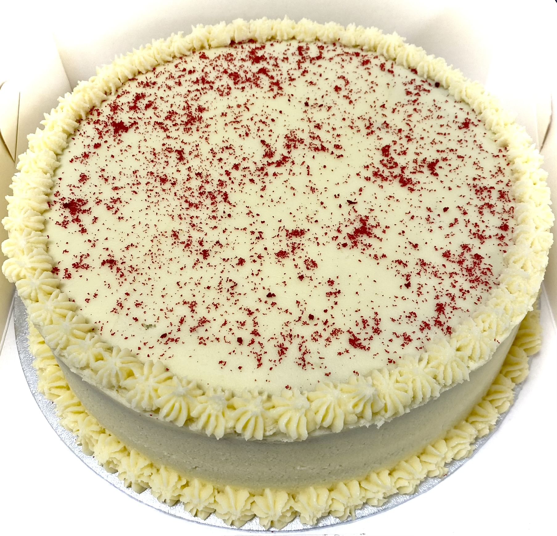A round red velvet cake with white cream cheese frosting. The top of the cake is sprinkled with red crumbs, and there is a decorative border of piped frosting along the edge at the base and top. The cake is placed on a silver cake board.